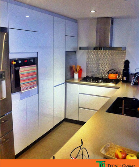 Cocina integral moderna cerrada sin manijas con pintura en poliuretano blanco. El meson es en quarztone caesarstone en tono beige. Se alcanza a ver parte de la nevera, el horno y encima del mesón utensilios y electrodomésticos