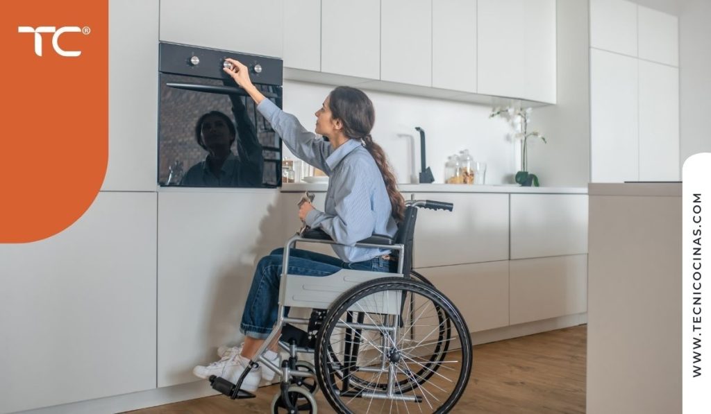 Mujer en silla de ruedas alcanzando el horno empotrado en una cocina con muebles blancos sin tirador